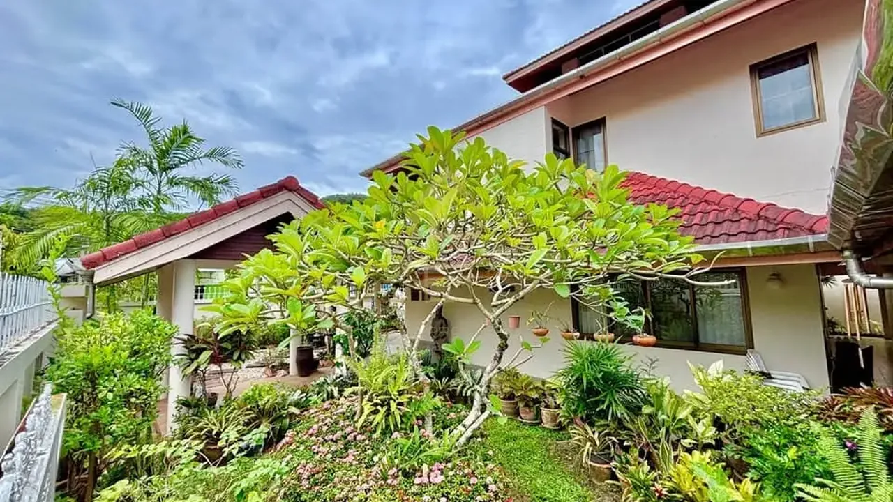 Ko Kaeo house: white two-storey villa with a pitched roof behind a lawn crossed by stepping stones, mature trees around it.