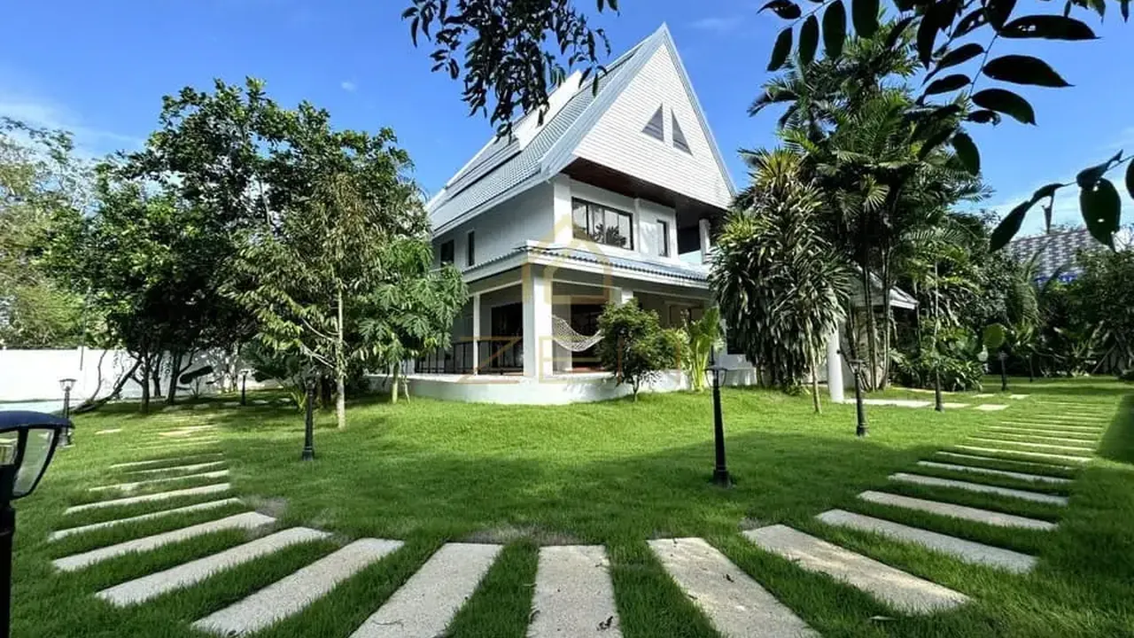 Pa Klok villa: white house with a steep gabled roof set back across a wide lawn with stepping stones and fruit trees.