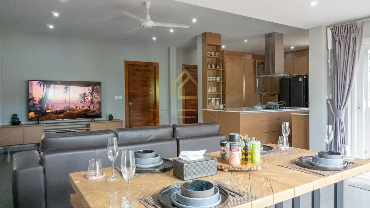Open-plan living and dining room with a long timber table set for dinner, a leather sofa and an island kitchen behind.