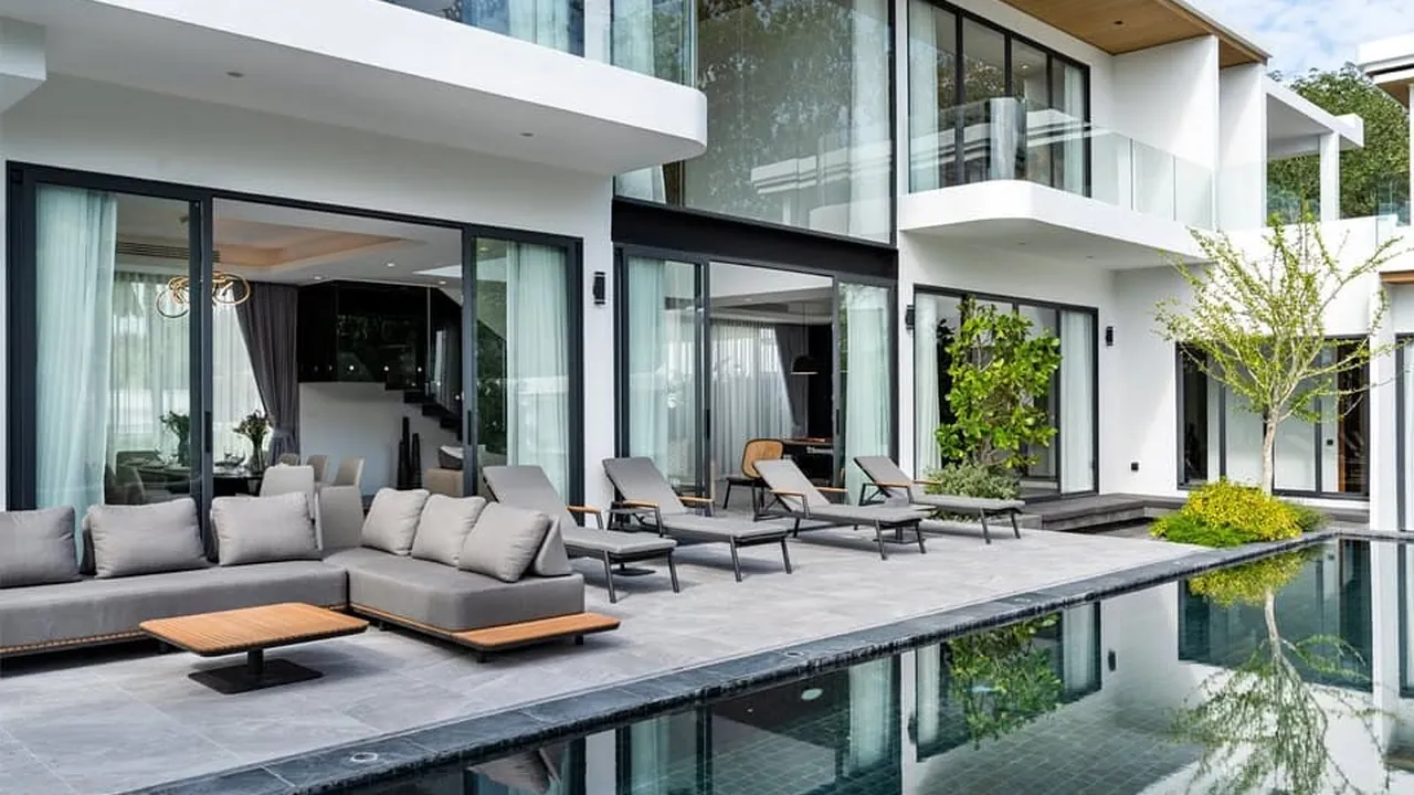 Four-bedroom Bang Tao villa: double-height white facade, grey outdoor sofas and loungers along a black-tiled pool.